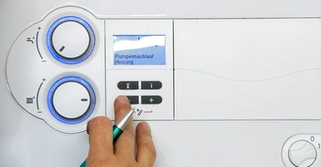 Eine Hand drückt Tasten neben einem Display auf einem weißen Bedienfeld mit zwei Drehreglern Pumpennachlauf Heizung