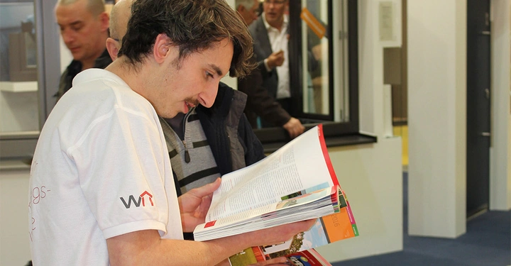 Galerie-Bild 3: Eine Person liest ein Magazin in einem Ausstellungsraum während zwei andere Männer im Hintergrund sprechen. Die Person trägt ein weißes T-Shirt mit dem Aufdruck WYT.