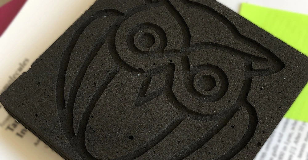 Schwarzer Schaumstoffblock zeigt das eingeprägte Bild einer Eule sitzt auf einer Unterlage bestehend aus verdecktem Text auf Papier und einem gelben Leuchtstiftpapier im Hintergrund.