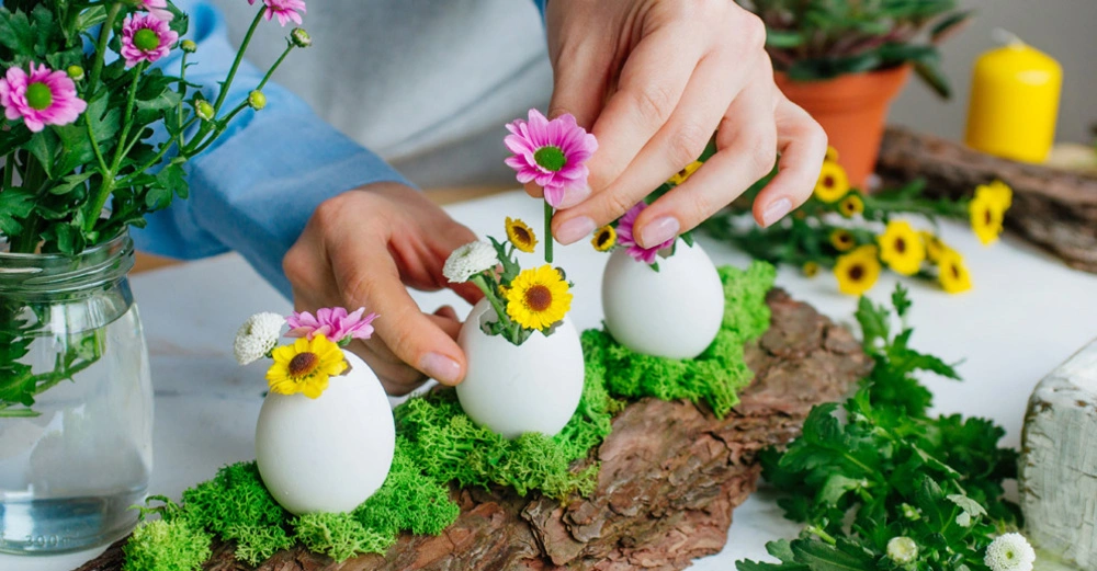 Frau dekoriert Eierschalen auf Holzbrettchen mit frischen Blüten und Kräutern, im Hintergrund gelbe Osterkerzen