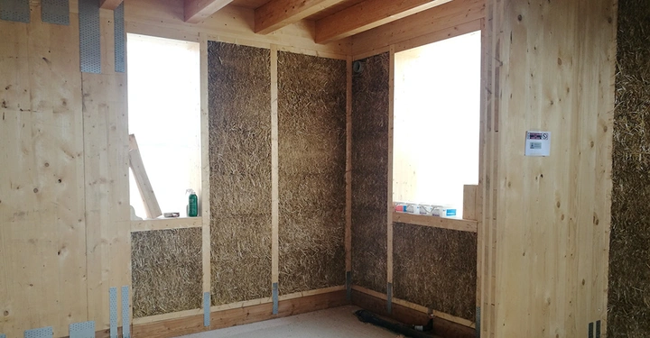 Innenraum mit unverkleideten Wänden aus Holz und Stroh. Bauarbeiten im Gang erkennbar durch Baumaterialien und Werkzeuge. Helle Atmosphäre durch Tageslicht von zwei Fenstern.