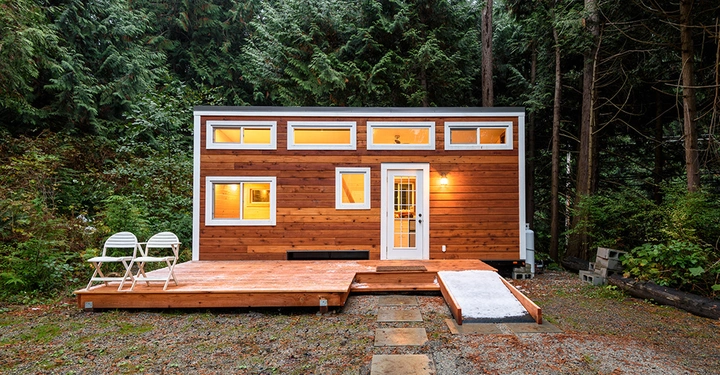Ein kleines Holzhaus mit mehreren Fenstern steht auf einer Lichtung im Wald. Zwei weiße Stühle und ein kleiner Tisch befinden sich auf der Holzterrasse vor dem Haus.