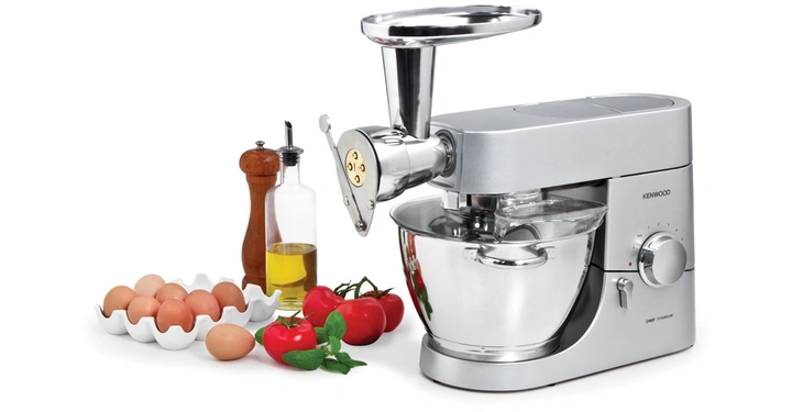 Küchenmaschine mischt Teig in einem silbernen Rührschüssel. Daneben stehen Eier, Tomaten, Basilikum, Olivenöl, Pfeffermühle. Umgebung ist eine saubere Küchenarbeitsfläche. Text auf der Maschine: KENWOOD CHEF TITANIUM.