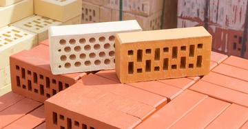 Zwei gelochte Ziegelsteine liegen auf gestapelten roten Ziegelsteinen im Freien, umgeben von Stapeln anderer Ziegelarten