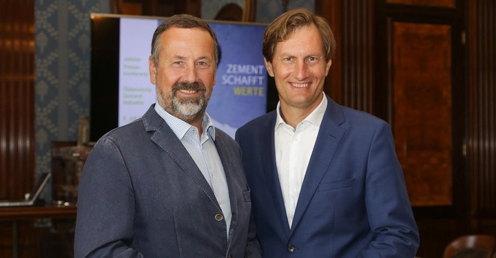 Zwei Männer stehen nebeneinander und lächeln in die Kamera. Sie befinden sich in einem Raum mit dunklen Holzverkleidungen. Text: ZEMENT SCHAFFT WERTE.