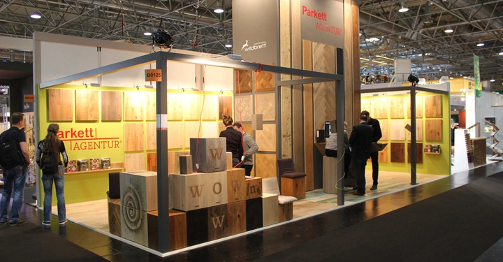 Galerie-Bild 16: Holzproben werden an einem Messestand namens Parkett Agentur präsentiert in einer Messehalle. Mehrere Personen betrachten die ausgestellten Holzprodukte und unterhalten sich miteinander. Text gefunden Parkett AGENTUR B0125 Wildbrett Parkett AGENTUR