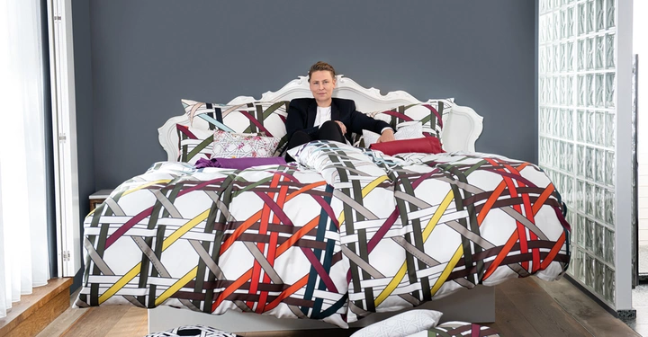 Person sitzt auf einem Bett. Das Bett hat eine weiße, gemusterte Bettwäsche. Das Zimmer hat dunkelgraue Wände, weiße Vorhänge und einen Holzboden und ist teilweise durch eine Glaswand abgetrennt.