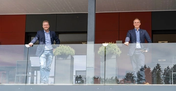 Zwei Männer lehnen sich an einem Glasgeländer auf einem Balkon. Beide tragen blaue Blazer und Jeans. Neben ihnen stehen Pflanzenbehälter. Der Hintergrund zeigt ein modernes Gebäude.