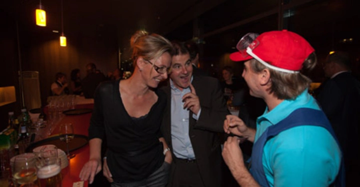 Galerie-Bild 81: Drei Personen unterhalten sich lächelnd an einer Bar Menschen und Getränke umgeben sie in einem gedämpft beleuchteten Raum mit moderner Ausstattung.