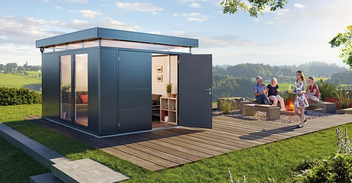 Grau-blaues Gartenhaus mit offener Tür steht auf Holzdeck Frau trägt Tablett Gruppe von Menschen sitzt auf Betonbank nahe Lagerfeuer grüner Rasen Wiesen und Bäume im Hintergrund.