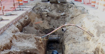 Ein Bagger gräbt einen tiefen Graben in einer Baustelle aus Straßenarbeiten mit Verkehrshütchen und Maschendrahtzäunen im Hintergrund. Rohre und Kies im Graben.