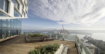 Eine Person sitzt auf einer Bank auf einer Dachterrasse mit Holzdecke an einem modernen Hochhaus Hafenbrücke und Opernhaus im Hintergrund bei bewölktem Himmel.