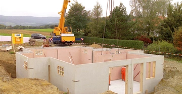 Ein Bauarbeiter richtet Betonteile im Rohbau eines Hauses aus ein Kran hebt Teile in eine Grube im Vordergrund Felder und ein Auto im Hintergrund Wald und Berge Text: KAMMEL Der Fertigkeller