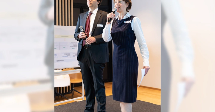 Galerie-Bild 2: Zwei Personen halten Mikrofone und sprechen auf einer Bühne in einem Konferenzraum. Eine Tafel mit Diagrammen und Text steht im Hintergrund auf einer Staffelei.