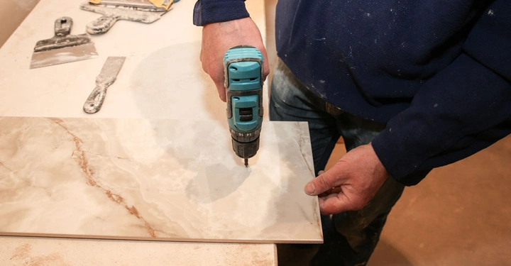 Person hält Bohrmaschine und bohrt in eine Fliese in einem Arbeitsbereich mit Werkzeugen auf einem Tisch.