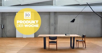 Ein Holztisch mit zwei Stühlen steht auf einem Holzfußboden in einem modernen Raum mit Betonwänden. Darüber ein großer runder Aufkleber mit der Aufschrift: 30 PRODUKT DER WOCHE
