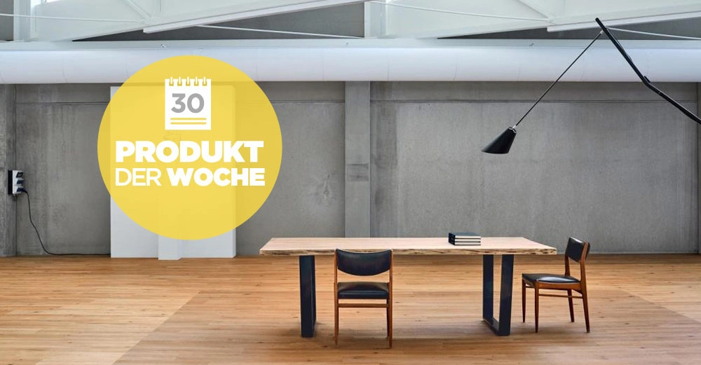 Ein Holztisch mit zwei Stühlen steht auf einem Holzfußboden in einem modernen Raum mit Betonwänden. Darüber ein großer runder Aufkleber mit der Aufschrift: 30 PRODUKT DER WOCHE