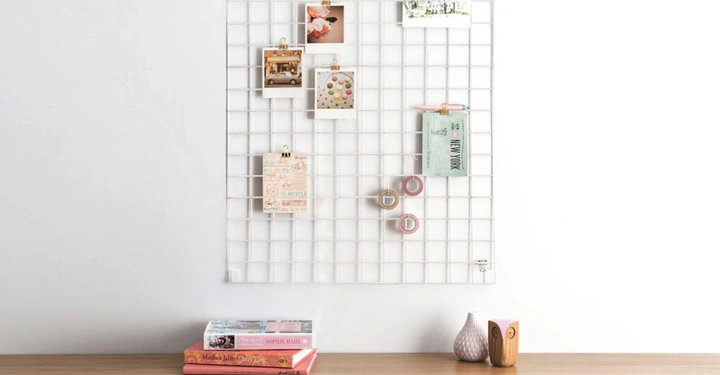 Ein weißes Gitter an einer Wand hält Fotos Notizen und Klebebänder darüber liegt eine Holzoberfläche mit mehreren Büchern und kleinen Vasen im Vordergrund