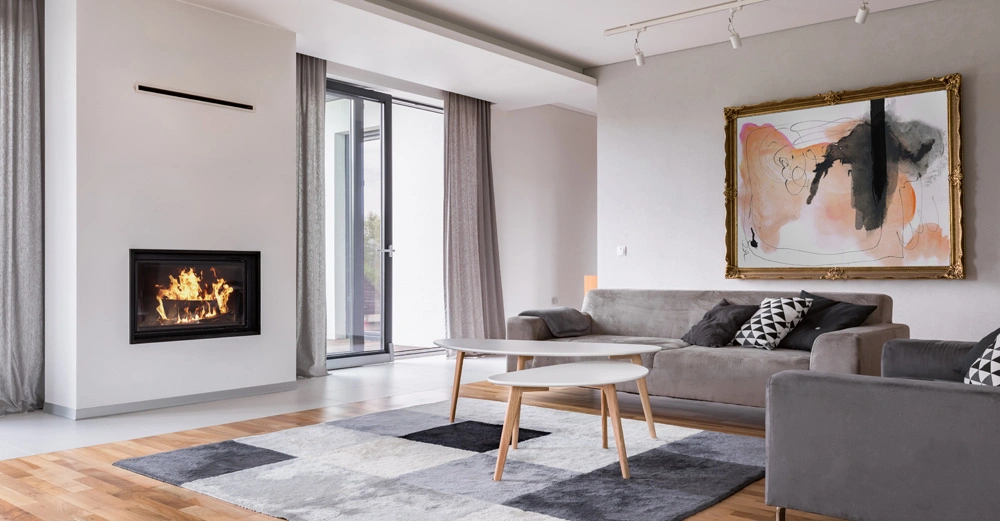 Kamin brennt Wohnzimmer moderne Einrichtung graue Sofas schwarz-weiße Kissen abstraktes Gemälde an der Wand grauer Teppich Holzfußboden große Fenster mit Vorhängen Couchtisch.