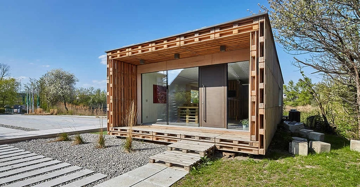Holzgebäude mit großen Glasfenstern steht auf Hölzern befindet sich in einer grünen Umgebung mit Bäumen und gemähtem Rasen unter klarem Himmel. Ein Weg führt zum Gebäude.