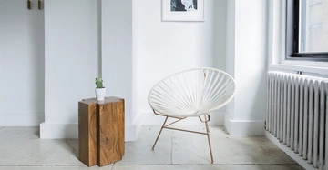 Ein weißer Stuhl steht auf einem hellen Boden neben einem kleinen Holztisch mit einem Kaktus darauf in einem minimalistischen Raum mit weiß gestrichenen Wänden und einem Heizkörper.