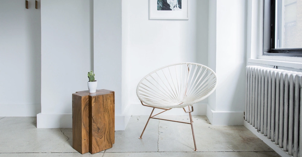 Ein weißer Stuhl steht auf einem hellen Boden neben einem kleinen Holztisch mit einem Kaktus darauf in einem minimalistischen Raum mit weiß gestrichenen Wänden und einem Heizkörper.
