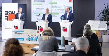 3 Männer im Anzug stehen bei Pressekonferenz auf Bühne an Stehpulten mit Baumit-Logo Aufdruck. Im Hintergrund ein großer Screen mit der Präsentation. Im Vordergrund Journalisten.