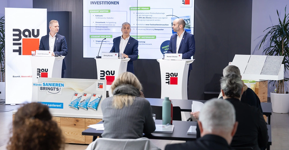 3 Männer im Anzug stehen bei Pressekonferenz auf Bühne an Stehpulten mit Baumit-Logo Aufdruck. Im Hintergrund ein großer Screen mit der Präsentation. Im Vordergrund Journalisten.
