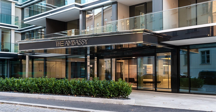 Galerie-Bild 2: Gebäude mit dem Schild THE AMBASSY vor dem Eingang Aufnahme der modernen Glas- und Metallfassade umgeben von Pflanzen und einer gepflasterten Straße