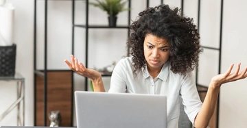 Eine Frau mit lockigem Haar hebt ratlos die Hände vor einem Laptop in einem modernen Büro mit Regalen und Pflanzen im Hintergrund