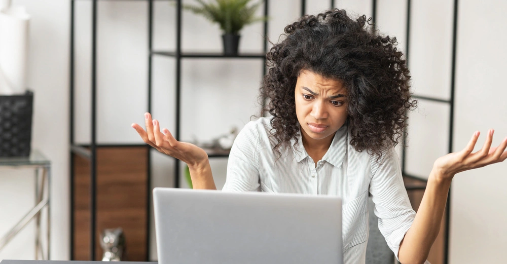 Eine Frau mit lockigem Haar hebt ratlos die Hände vor einem Laptop in einem modernen Büro mit Regalen und Pflanzen im Hintergrund