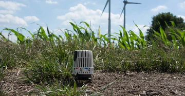 Ein Thermostatkopf steht im Vordergrund auf einer Wiese mit Gras und Pflanzen umgeben zwei Windräder stehen im Hintergrund unter einem blauen Himmel mit Wolken. Der Thermostatkopf zeigt die Zahlen 2 3 4.