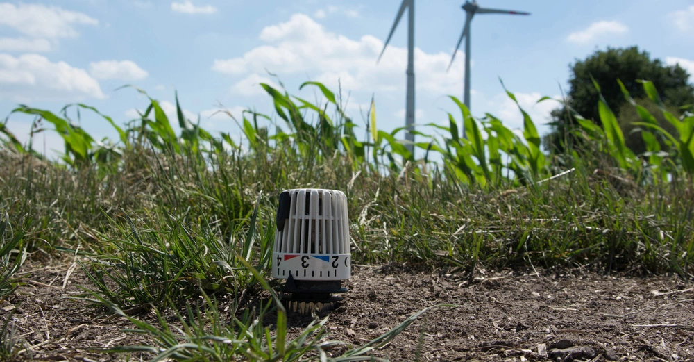 Ein Thermostatkopf steht im Vordergrund auf einer Wiese mit Gras und Pflanzen umgeben zwei Windräder stehen im Hintergrund unter einem blauen Himmel mit Wolken. Der Thermostatkopf zeigt die Zahlen 2 3 4.