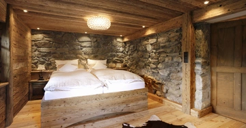 Ein rustikales Schlafzimmer zeigt ein Doppelbett mit karierter Bettwäsche vor groben Steinwänden und Holzwänden. Im Vordergrund liegt ein Kuhfell-Teppich auf hellem Holzfußboden.