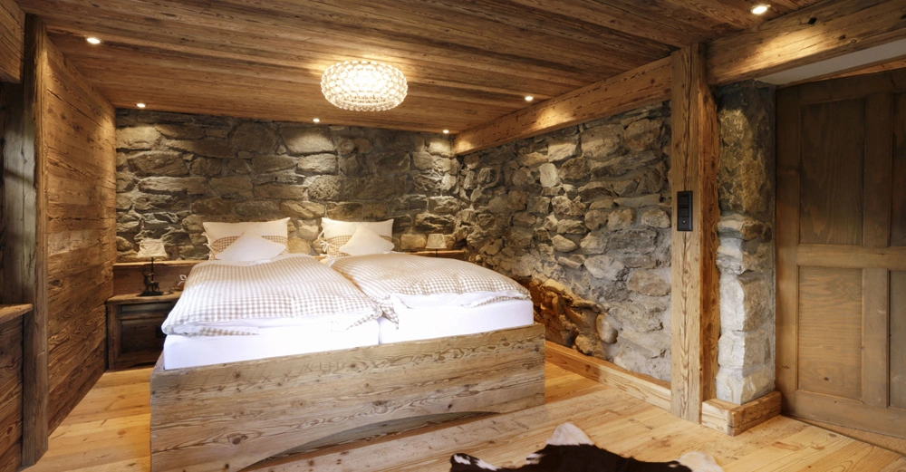 Ein rustikales Schlafzimmer zeigt ein Doppelbett mit karierter Bettwäsche vor groben Steinwänden und Holzwänden. Im Vordergrund liegt ein Kuhfell-Teppich auf hellem Holzfußboden.