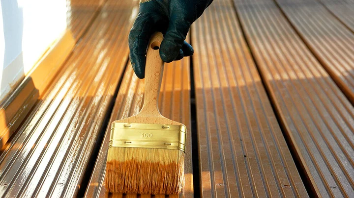 Hand mit schwarzem Handschuh hält großen Pinsel und ölt Terrassenboden ein