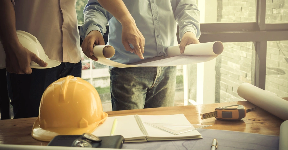 Zwei Personen betrachten Baupläne in einem Büro. Auf einem Tisch liegen ein gelber Bauhelm, ein Notizbuch, ein Maßband und weitere Bauzeichnungen. Hintergrund zeigt eine große Fensterfront.