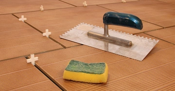 Verputzkelle und gelb-grüner Schwamm liegen auf frisch verfliestem Cotto-Boden. Zwischen den Fliesen stecken Fugenkreuze aus Kunststoff.