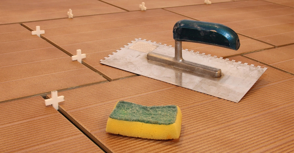 Verputzkelle und gelb-grüner Schwamm liegen auf frisch verfliestem Cotto-Boden. Zwischen den Fliesen stecken Fugenkreuze aus Kunststoff.
