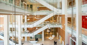 Ein mehrstöckiges modernes Gebäude Auditorium mit nummerierten Treppen (1-4) führt zu offenen Gängen mit Menschen, die sich auf verschiedenen Etagen bewegen. Helle und minimalistische Inneneinrichtung umgeben die Szene.