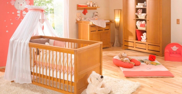 Galerie-Bild 5: Babybett mit weißem Baldachin steht auf einem Teppich Teddybär davor Kommode und Schrank im Hintergrund andere Kuscheltiere und Spielzeug auf Spielmatte Holzboden mit rot weißen Wänden und Wandstickern