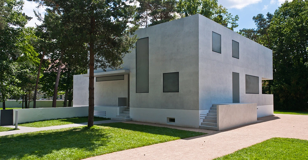Modernes minimalistisches Einfamilienhaus mit klarer kubischer Architektur und grauer Betonfassade in grüner Umgebung.