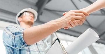 Zwei Personen geben sich die Hand einer trägt einen Schutzhelm und hält Baupläne, im Hintergrund eine moderne Gebäudestruktur.