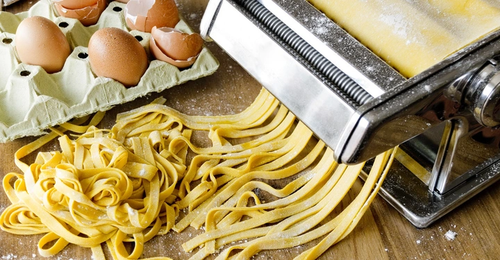 Nudelmaschine produziert frische Nudeln auf hölzernem Küchentisch neben Eierschachtel mit Eiern und Eierschalen in einer bemehlten Umgebung.