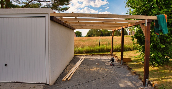 Eine teilweise gebaute Carportkonstruktion steht neben einer weißen Garage mit heruntergelassener Tür in einer ländlichen Umgebung mit Bäumen und Feldern im Hintergrund