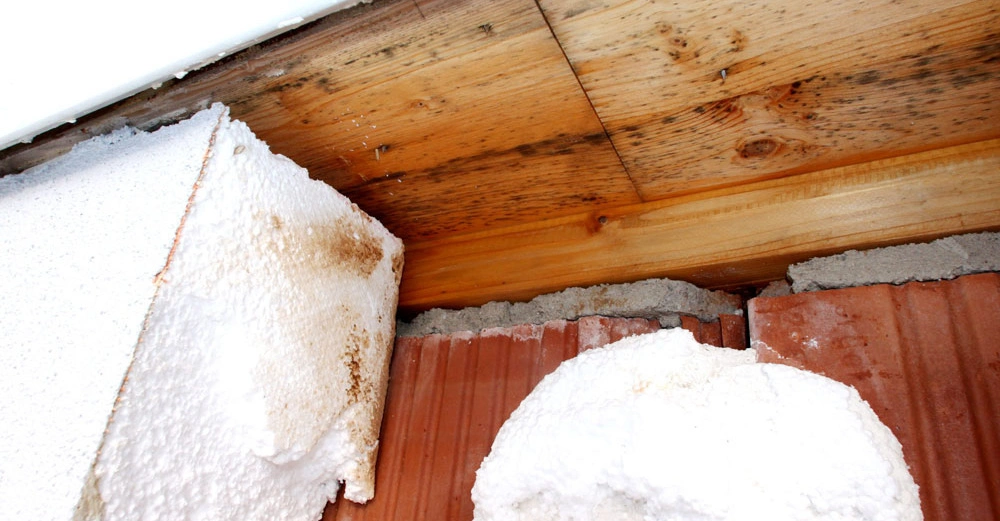 Styroporplatten liegen schief an einer Holzdecke in einem unvollendeten Bauprojekt an. Die Umgebung zeigt unverputzte Ziegel und einige Stellen auf dem Styropor sind verfärbt.