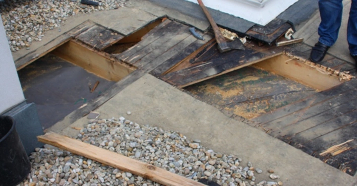 Galerie-Bild 8: Eine beschädigte Holzplattform wird mit Werkzeugen wie einer Axt und einem Brecheisen bearbeitet. Die Umgebung besteht aus einem Kiesdach mit größeren Flächen freigelegten Holzes und Wasseransammlungen.