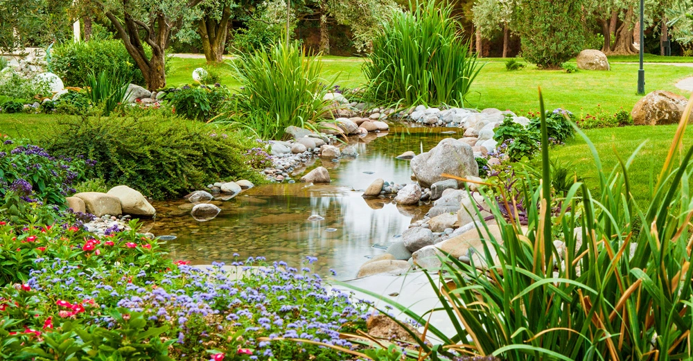 Bach fließt durch gepflegten Garten mit bunten Blumen und grünen Sträuchern unter Bäumen.