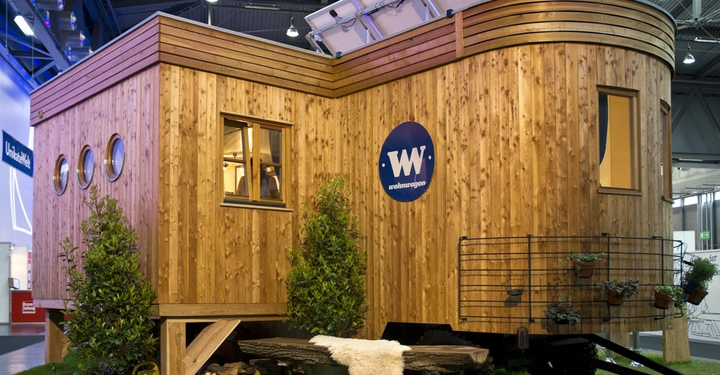 Galerie-Bild 2: Ein Holzwagen auf Rädern steht auf einer grünen Rasenfläche in einer überdachten Innenausstellung mit Metallträgern. Text auf dem Wagen WW wohnwagon