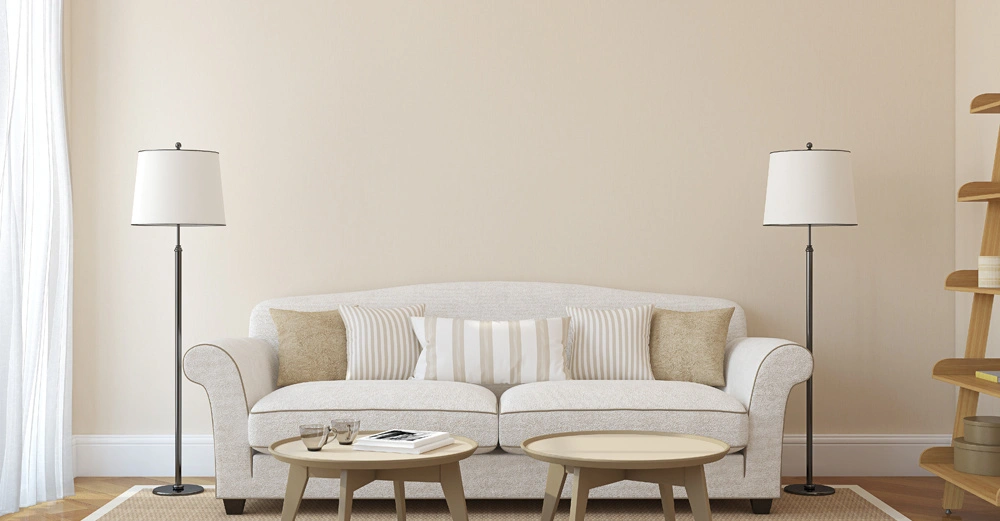 Ein beige Sofa steht zwischen zwei Stehlampen in einem hellen Wohnzimmer, umgeben von einem Teppich und zwei Couchtischen mit Gläsern und Büchern. Rechts steht ein Regal.